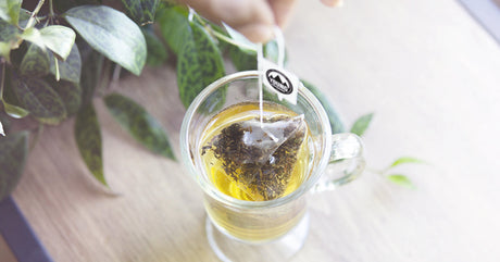 A pyramid tea bag being dipped into a glass mug filled with hot water.