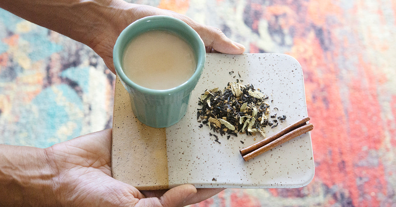 Hands holding a ceramic tray, carrying a filled chai cup, loose-leaf chai blend and cinnamon stick.