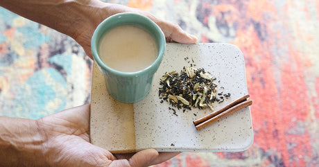 Hands holding a ceramic tray, carrying a filled chai cup, loose-leaf chai blend and cinnamon stick.